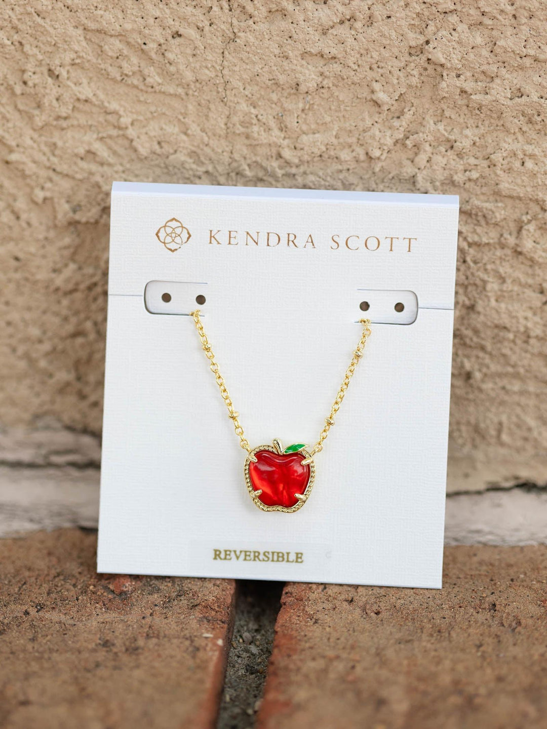 A gold chain necklace with a red apple pendant, displayed on a white Kendra Scott card against a textured beige wall.
