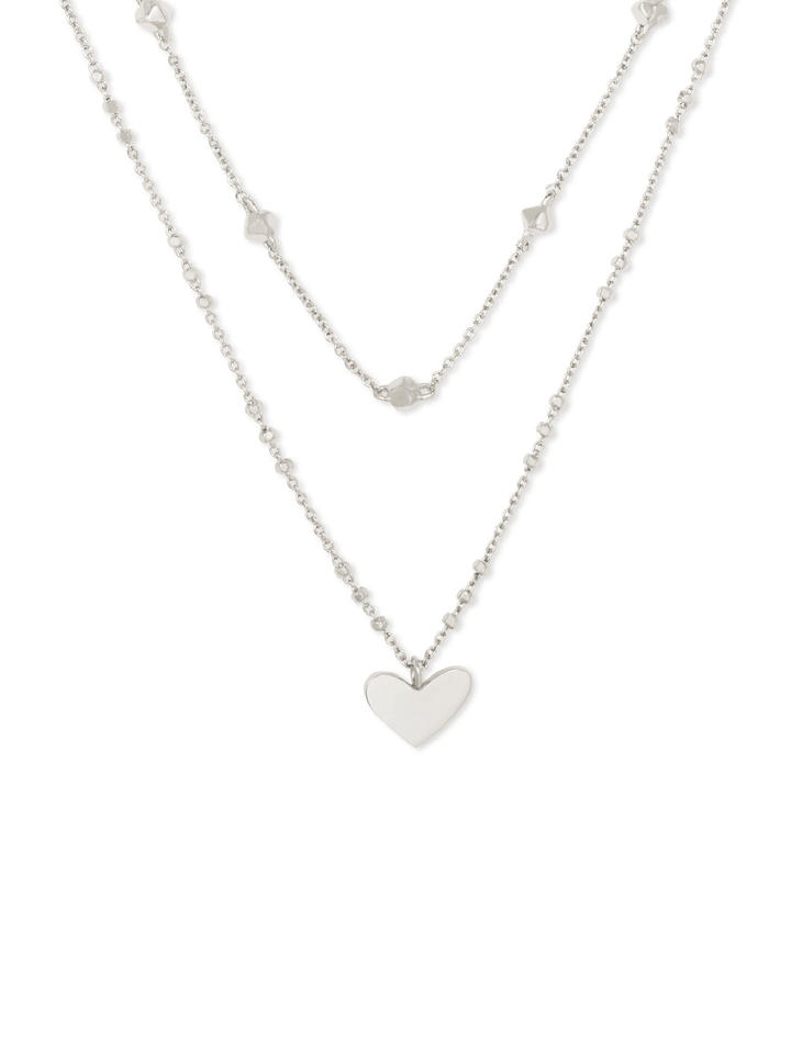 A silver double-layered necklace featuring a delicate chain with small star-shaped charms and a longer chain with a polished heart pendant, displayed against a solid white background.