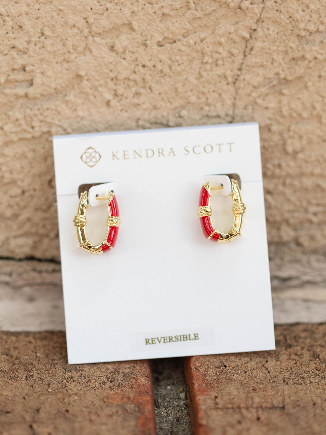 A pair of small gold hoop earrings with red inlay, displayed on a white Kendra Scott card against a textured beige wall.