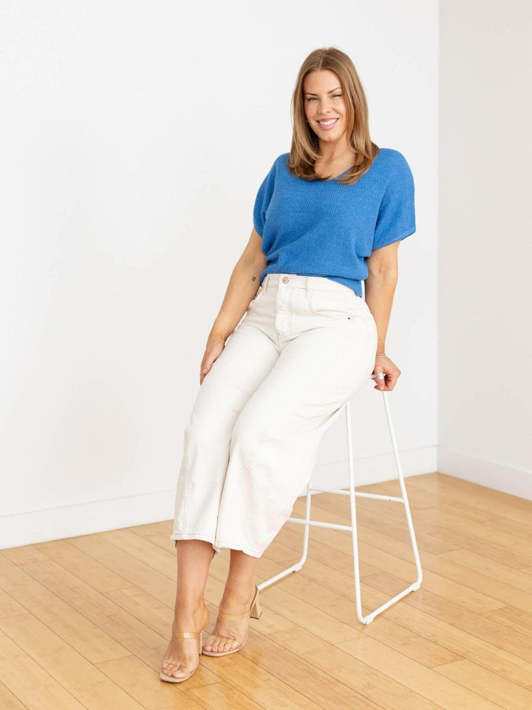 Woman in a blue top and white pants standing in a minimalistic room with a white wall and wooden floor.