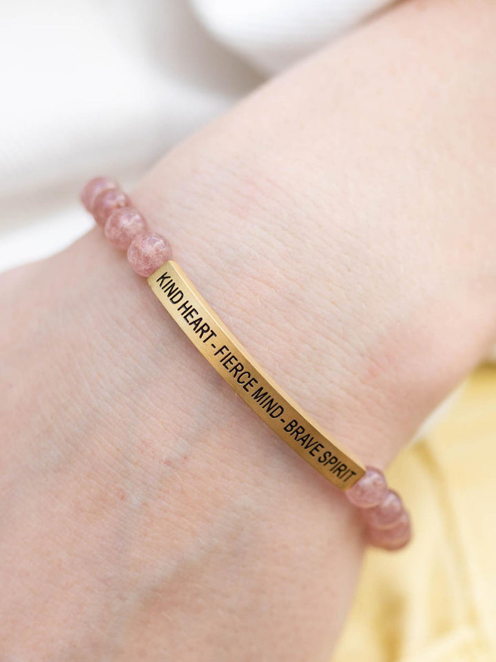 Pink Quartz stone bracelet with a gold bar engraved "KIND HEART - FIERCE MIND - BRAVE SPIRIT" on a wrist.
