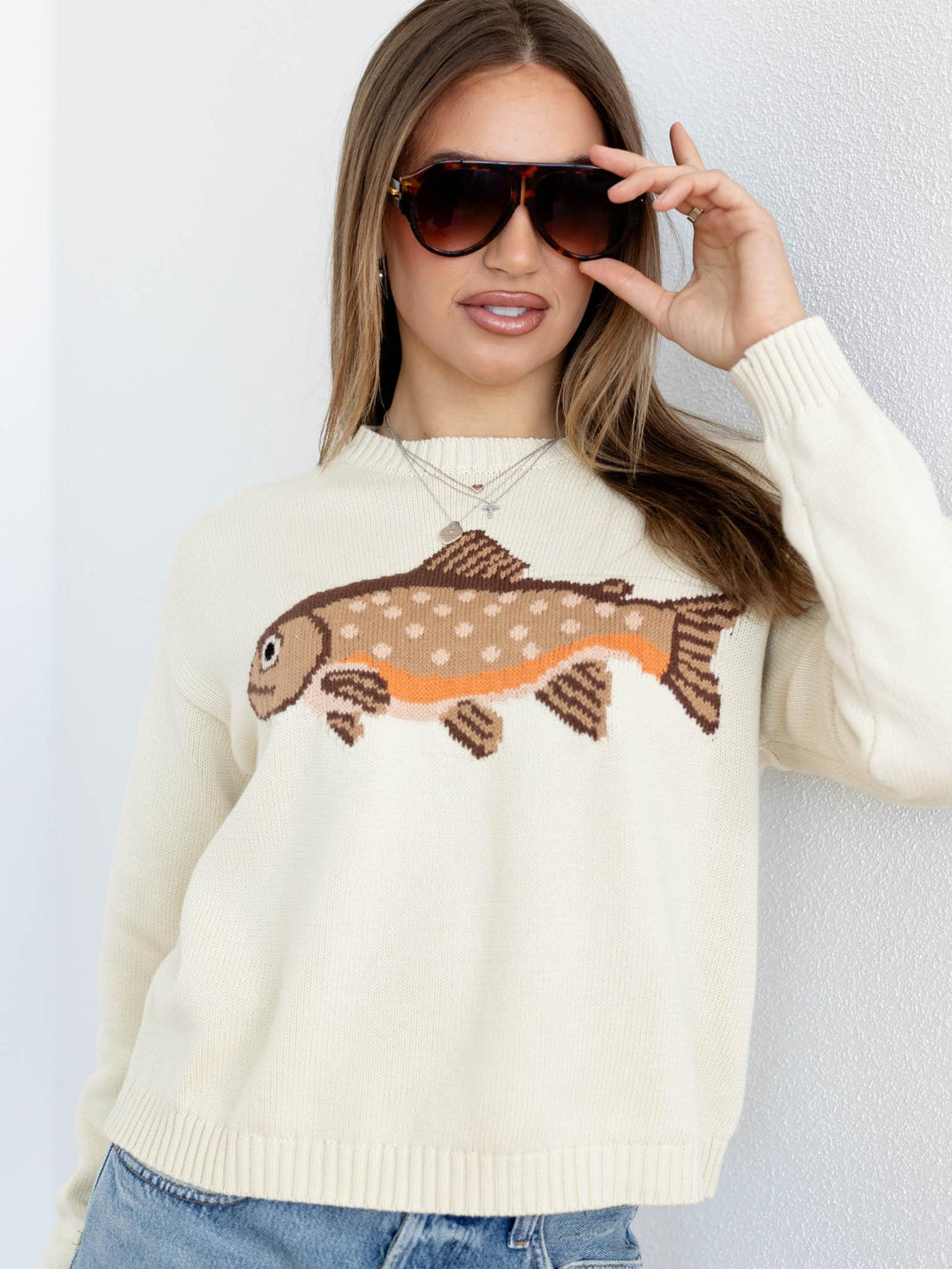 Woman wearing a cream sweater with a fish design, sunglasses, and blue jeans against a light gray background.
