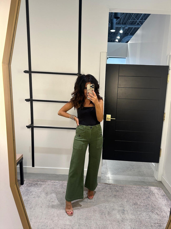 Person taking a mirror selfie wearing a black top and green pants in a room with a black door and ladder.