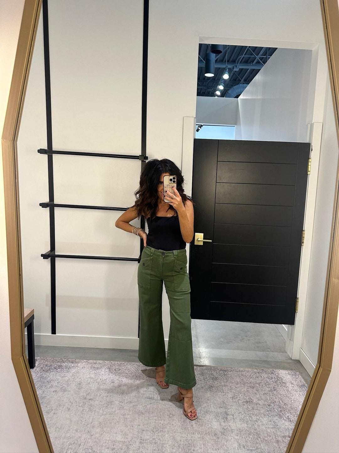 Person taking a mirror selfie wearing a black top and green pants in a room with a black door and white walls.