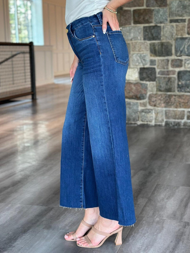 A person wearing blue wide-leg cropped jeans with a frayed hem, paired with beige heeled sandals, standing in an indoor space with a stone wall and wood floor.