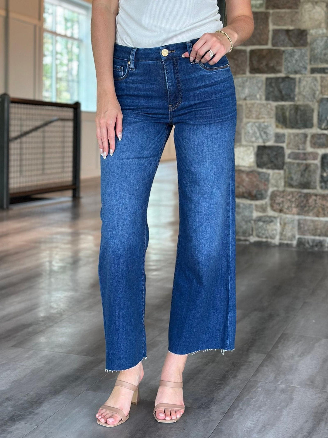 A person wearing medium-wash blue cropped flare jeans with a raw hem, paired with a white top and beige heeled sandals, standing in an indoor space with a stone wall and wood floor.