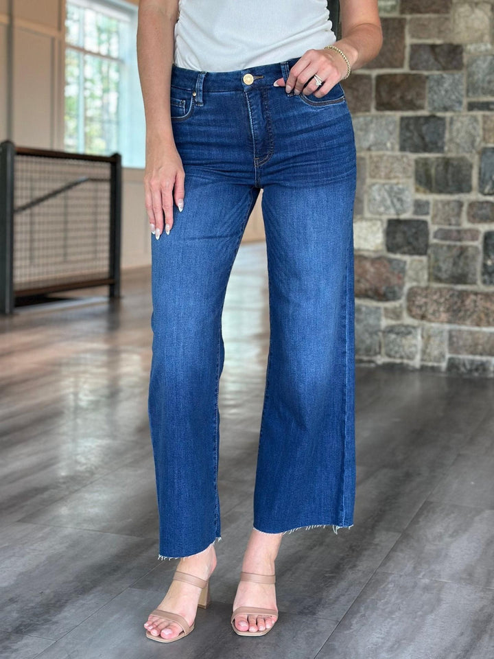 A person wearing medium-wash blue cropped flare jeans with a raw hem, paired with a white top and beige heeled sandals, standing in an indoor space with a stone wall and wood floor.