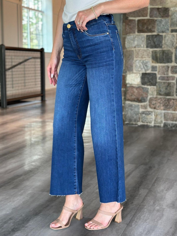 A person wearing high-waisted, wide-leg blue denim jeans with a raw hem, paired with beige heeled sandals in an indoor setting with a stone wall and wood floor.