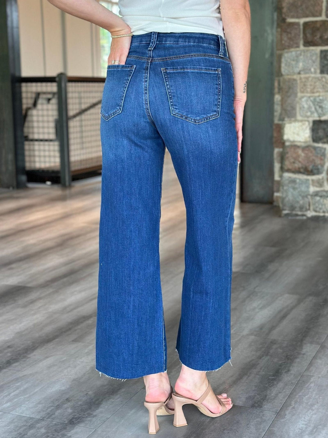 A person shown from the back wearing blue wide-leg cropped jeans with a frayed hem, paired with a white top and beige heeled sandals in an indoor setting with wood flooring and a stone wall.