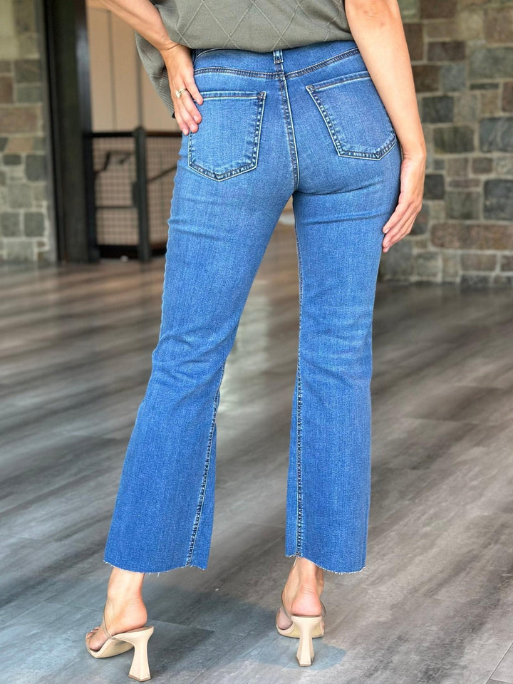 A person shown from the back wearing blue flare jeans with side slits, paired with a gray top and beige heeled sandals, standing on a wood floor in front of a stone wall.