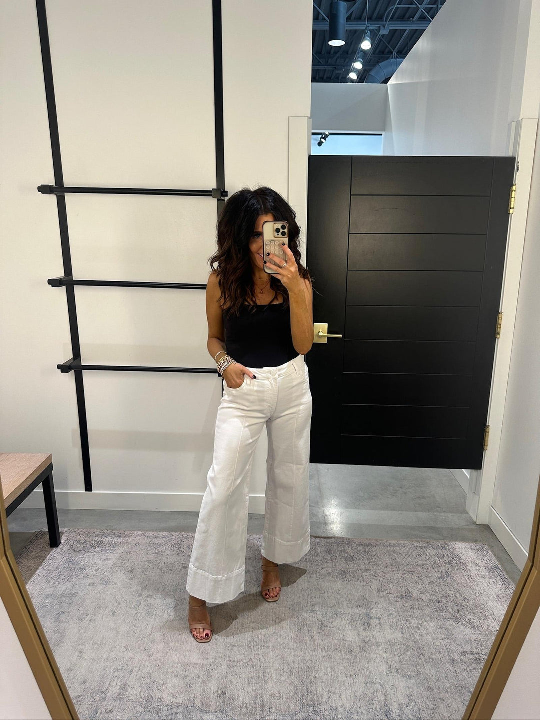 Woman taking a mirror selfie wearing a black top and white pants in a room with a black wall and gray carpet.