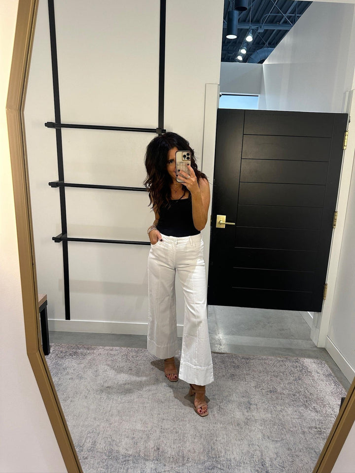 Woman taking a mirror selfie wearing a black top and white pants in a room with a black door and gold ladder.