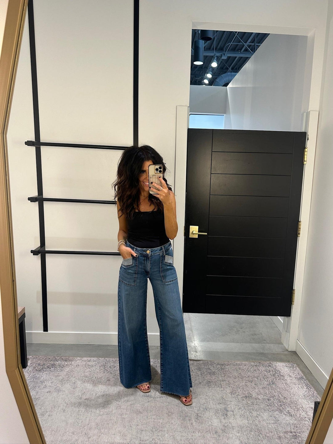 Person taking a mirror selfie wearing a black top and blue jeans in a room with a black door and ladder.