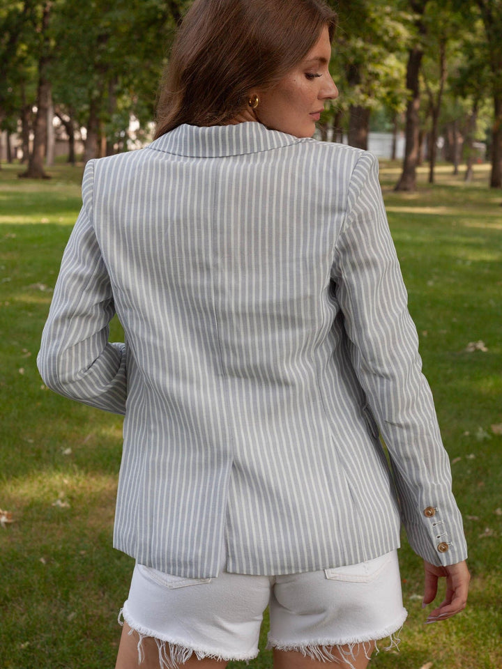 A woman viewed from behind wearing a light gray and white vertically striped blazer with white frayed denim shorts, standing in a grassy park with trees in the background.