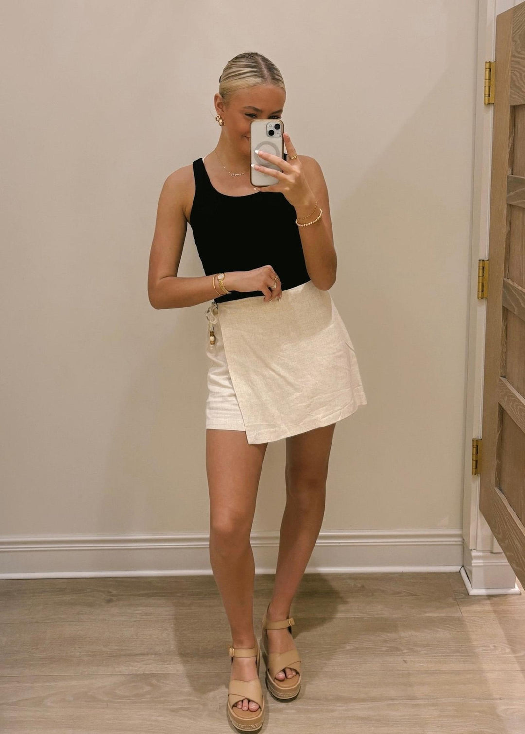 Person wearing a black tank top and beige skirt taking a mirror selfie.