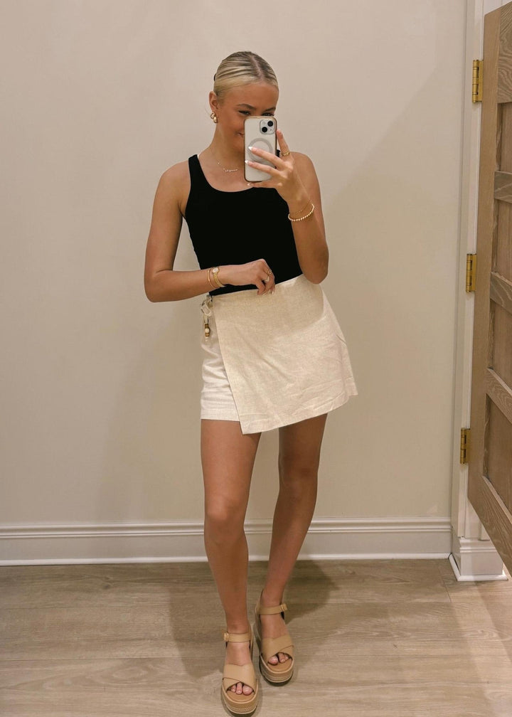 Person wearing a black tank top and beige skirt taking a mirror selfie.