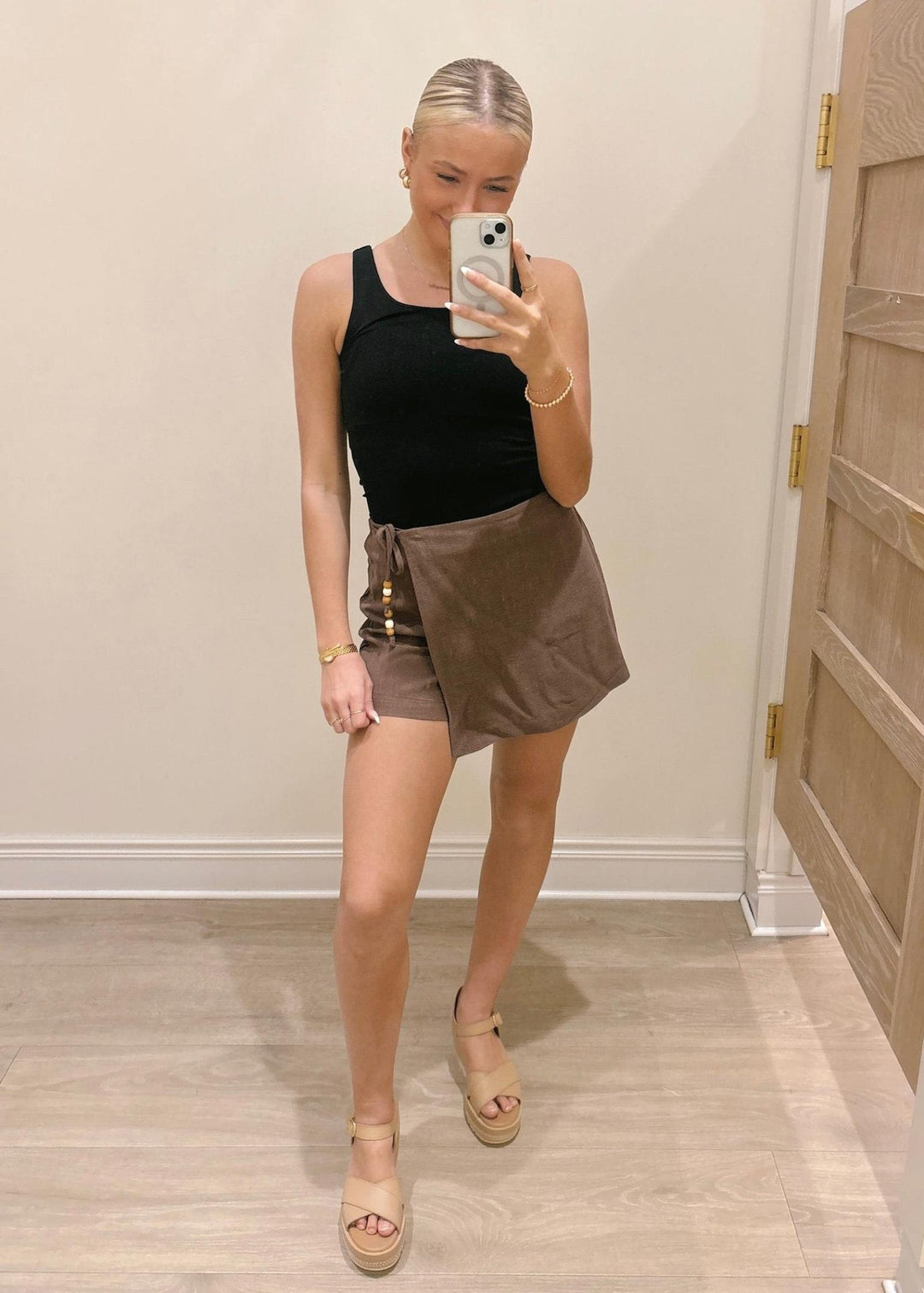 Woman taking a mirror selfie wearing a black tank top and brown shorts in a room.
