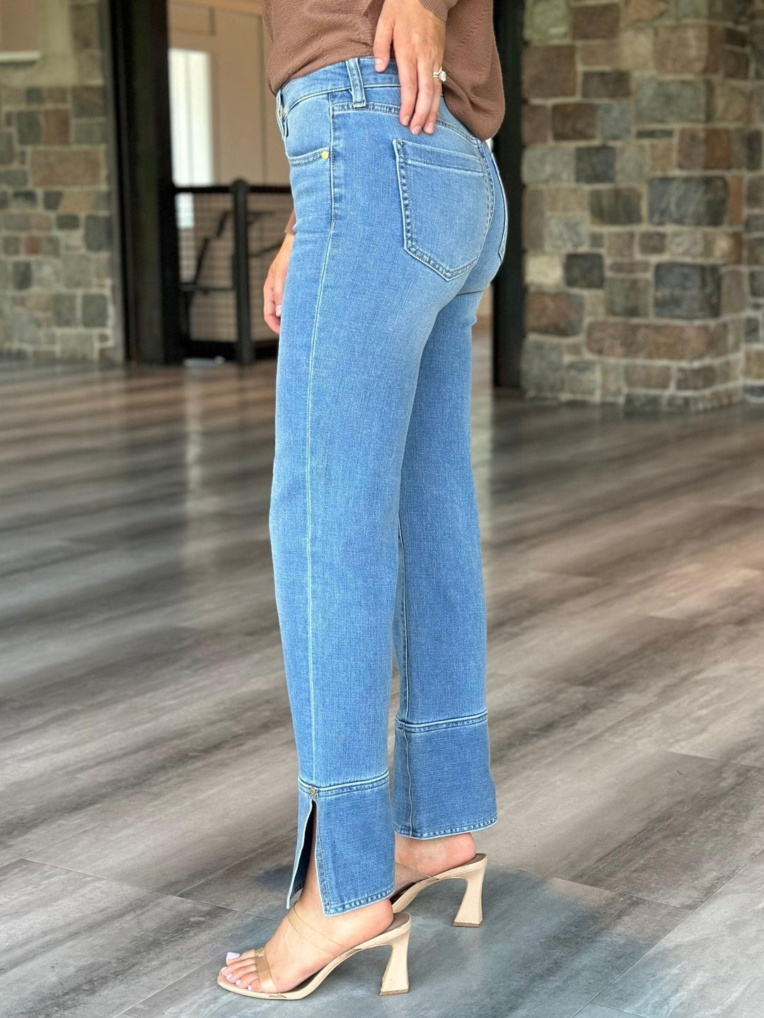 A person wearing light blue cropped jeans with side slits and a brown top, shown from the back in an indoor setting with wood flooring and stone walls.