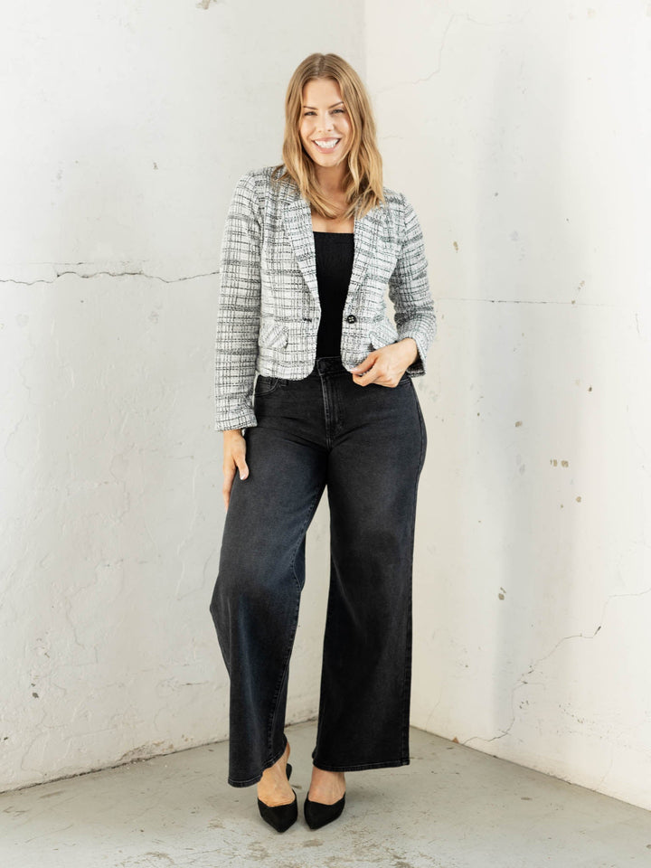 Woman wearing a plaid jacket, black top, and wide-leg jeans against a white brick wall.