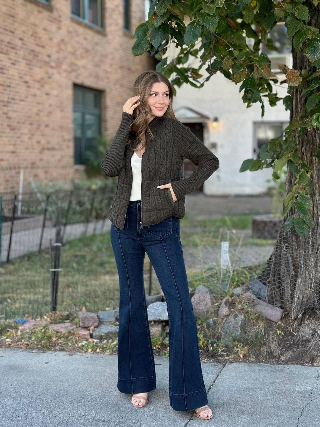 A woman stands outdoors on a sidewalk wearing a dark olive green cable-knit zip-front cardigan over a white top, paired with dark blue flared jeans and pink sandals. A brick building, grass, and a tree are visible in the background.