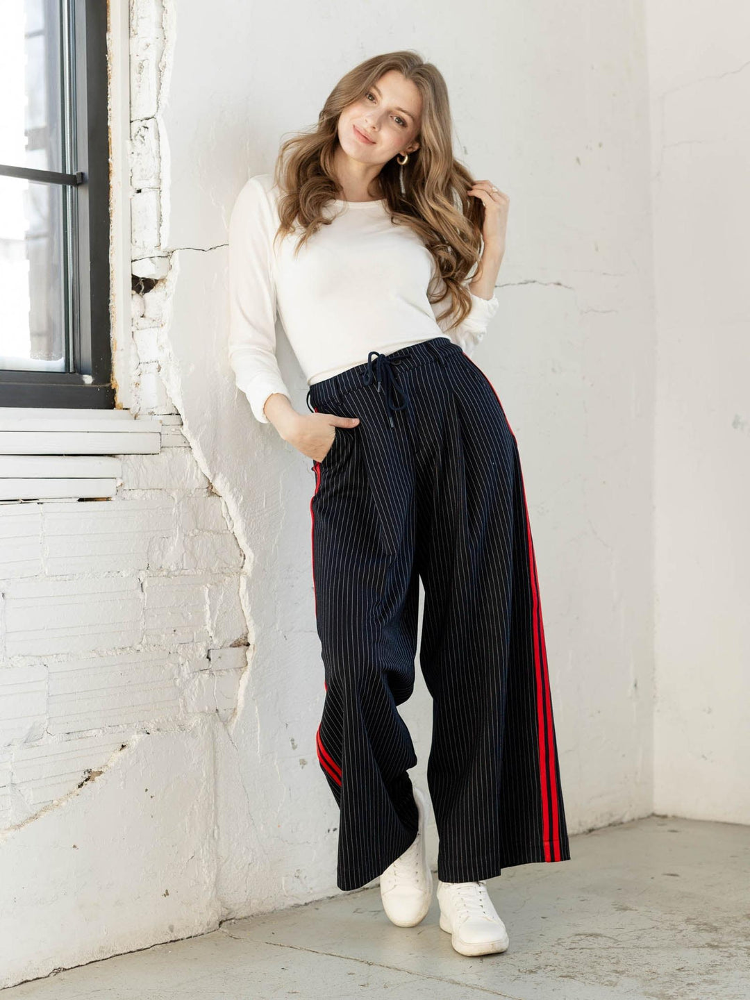 Woman wearing a white top and black pants with red stripes, standing against a white brick wall.