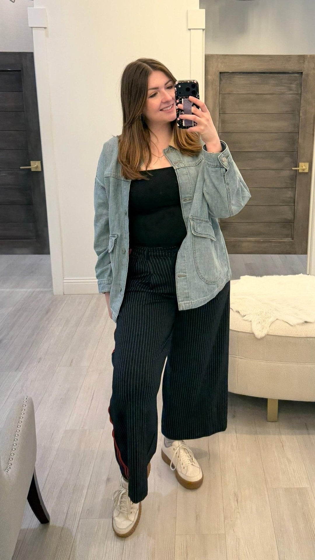 Woman taking a mirror selfie wearing a light blue denim jacket, black top, and dark pants in a room.