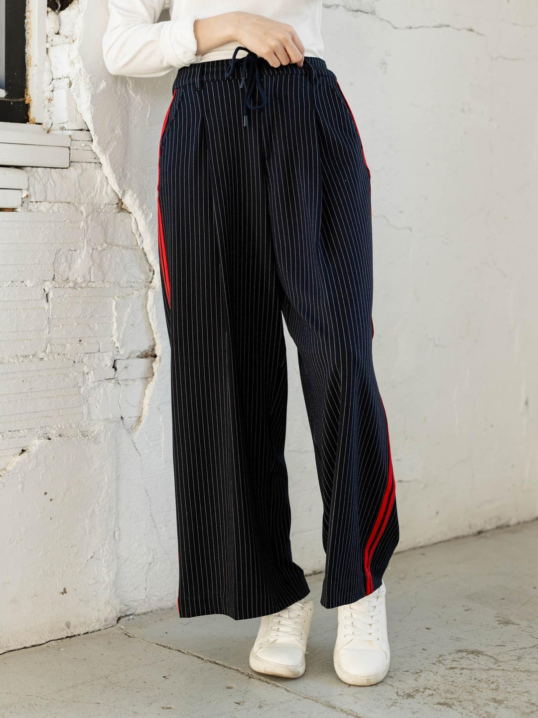 Person wearing black pleated pants with red stripes against a white brick wall.