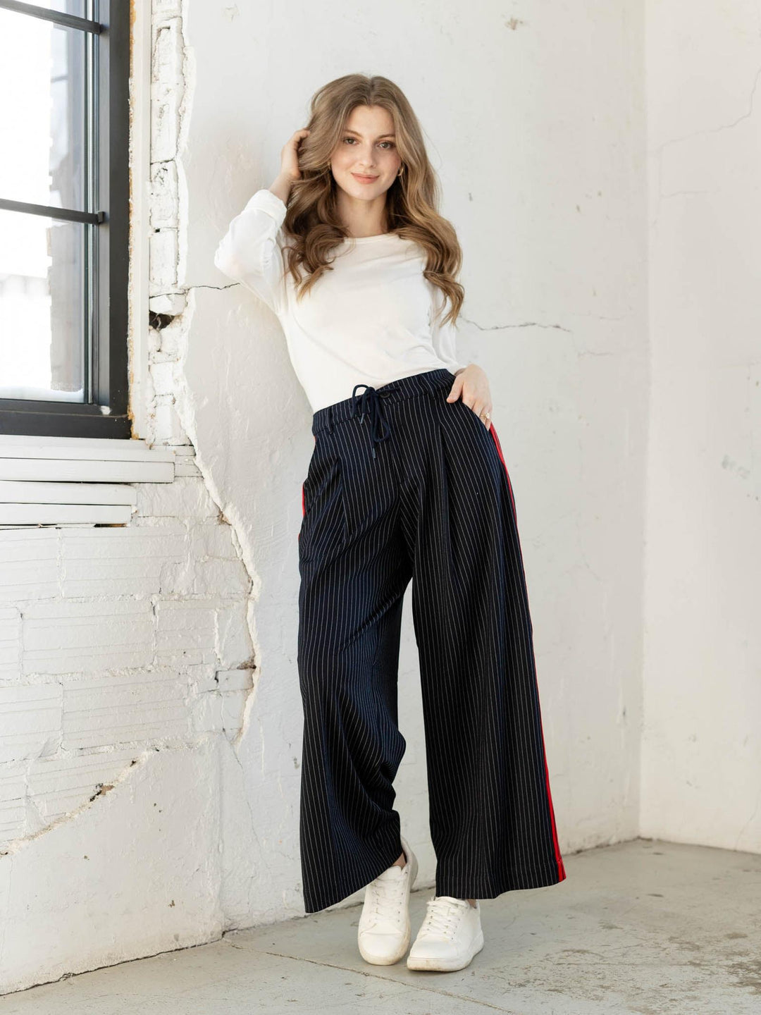 Woman wearing a white sweater and black pants with red stripes, standing against a white brick wall.