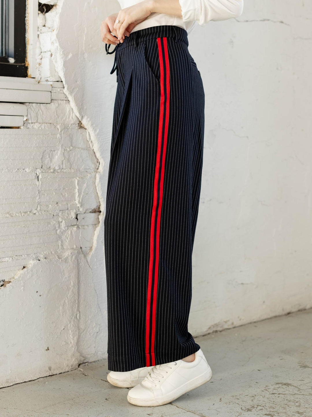 Person wearing black pleated pants with red stripes against a white wall.