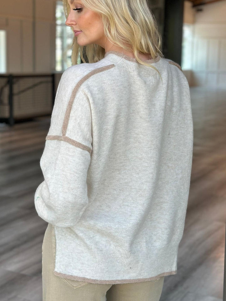 A woman wearing a light beige knit sweater with contrasting tan trim details on the shoulders and sleeves, shown from a side-back angle in an indoor setting.
