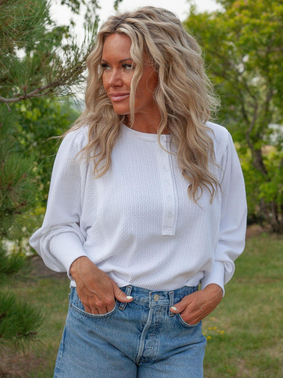 A woman wearing a white long-sleeve waffle-knit top with a buttoned henley collar and light blue jeans, standing outdoors in a grassy area with trees in the background.