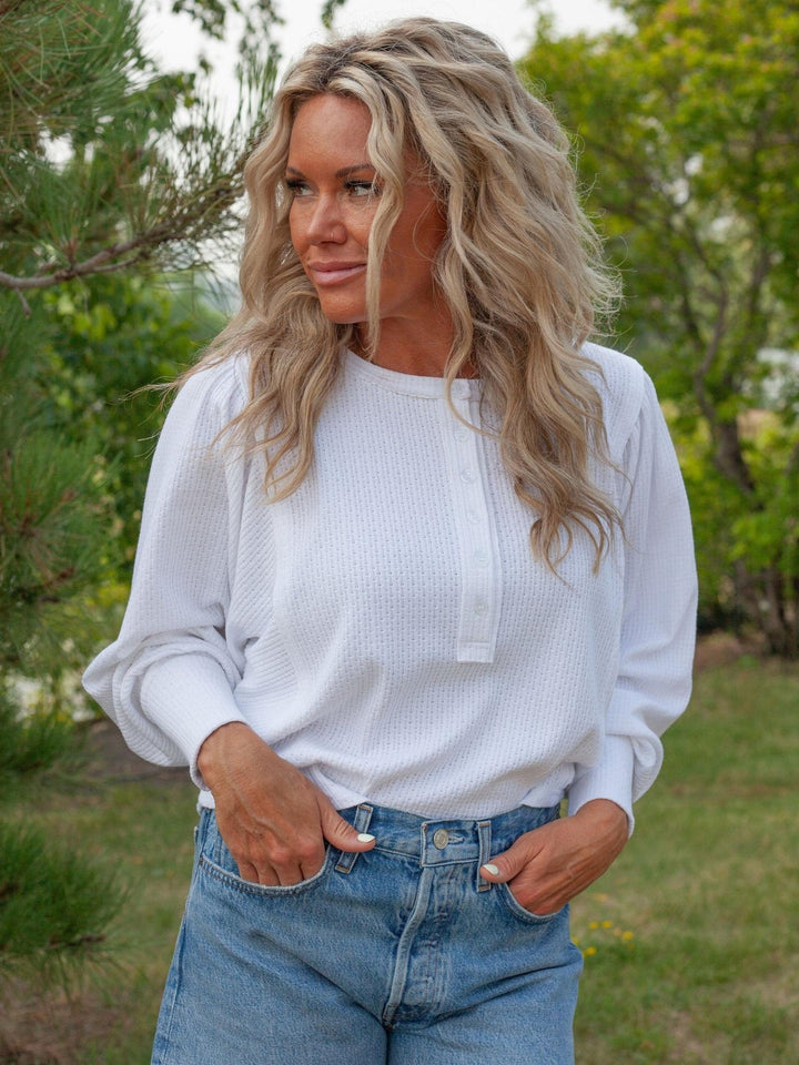 A woman wearing a white long-sleeve waffle-knit top with a buttoned henley collar and light blue jeans, standing outdoors in a grassy area with trees in the background.