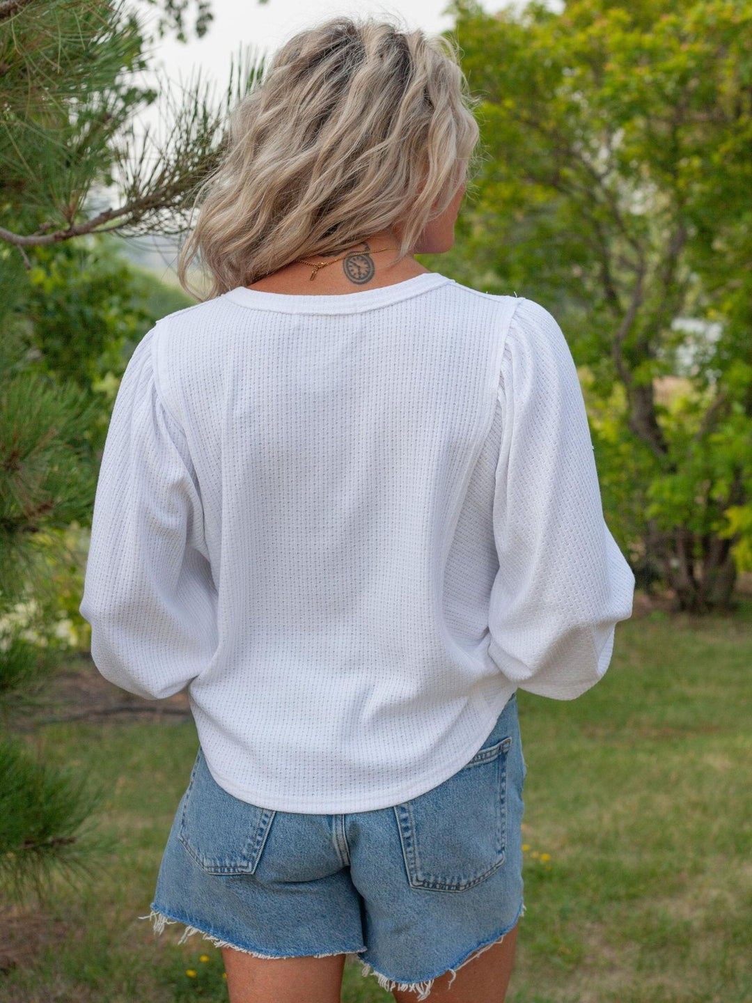A woman viewed from behind wearing a white textured long-sleeve top and denim shorts in an outdoor grassy setting with trees.
