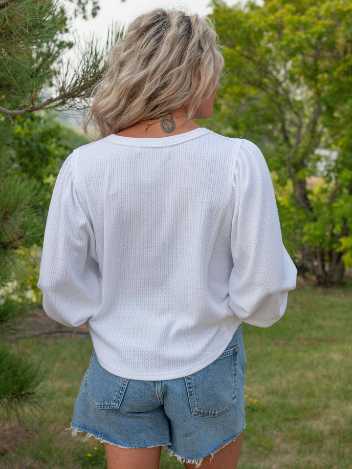 A woman viewed from behind wearing a white textured long-sleeve top and denim shorts in an outdoor grassy setting with trees.