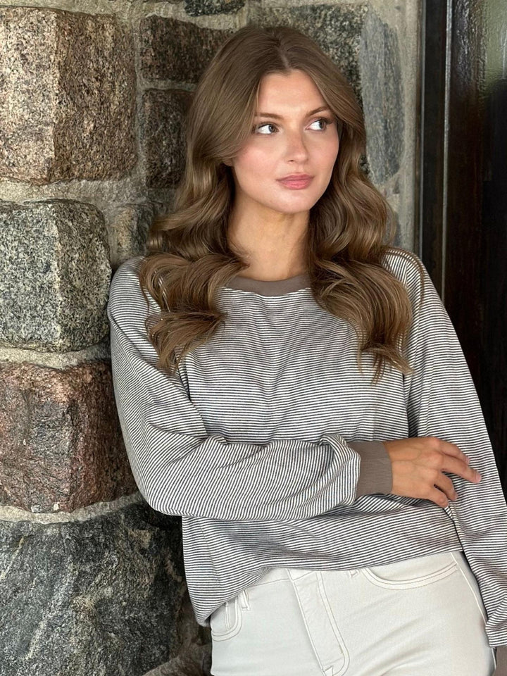 A woman with long wavy brown hair leans against a rustic stone wall, wearing a gray and white horizontally striped long-sleeve crewneck sweatshirt and white pants.