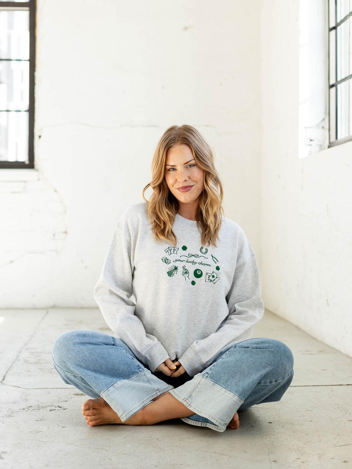Woman sitting cross-legged on a white floor wearing a light gray sweatshirt with green text and blue jeans.