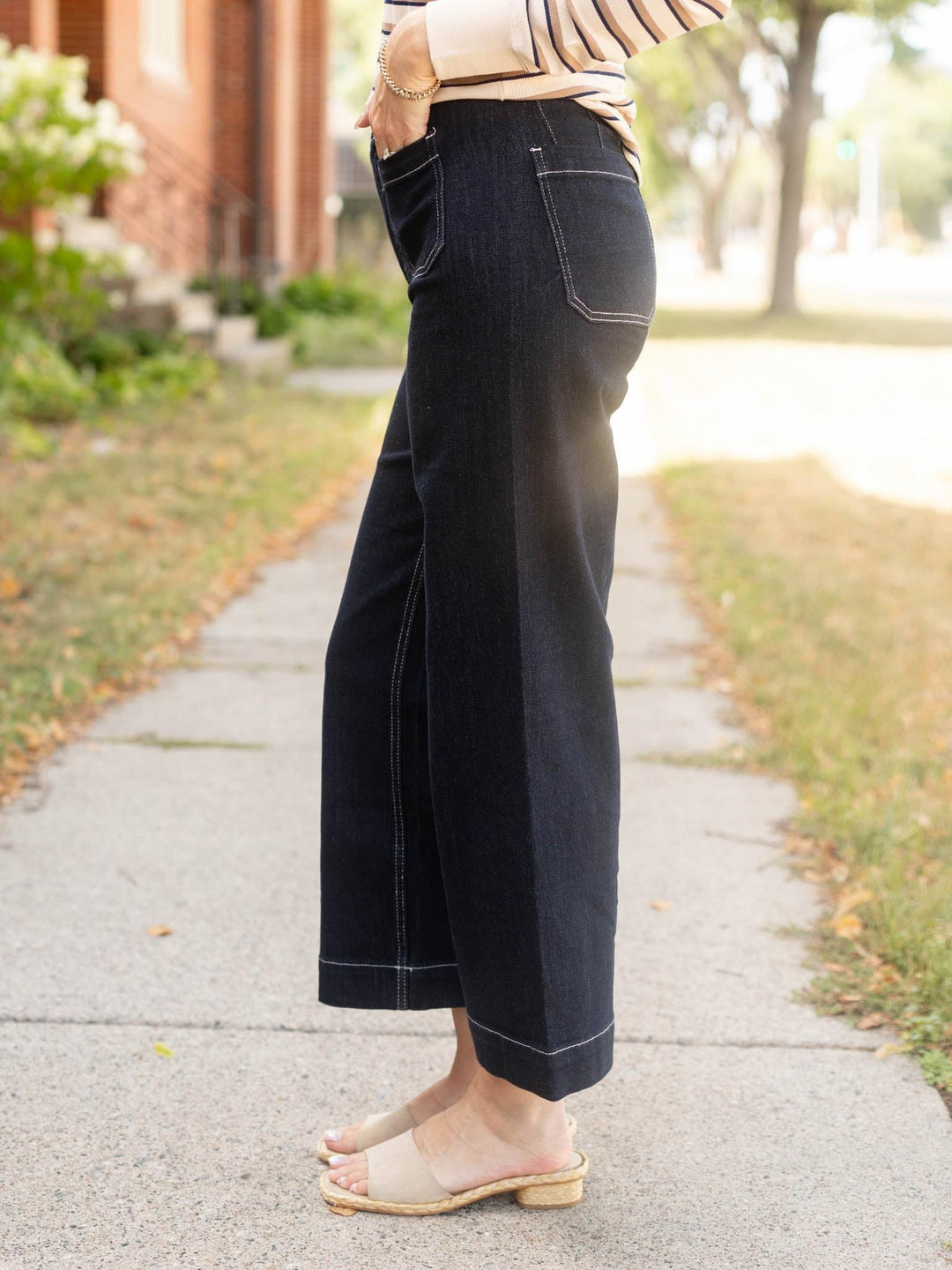 A person wearing dark wide-leg cropped jeans with contrast light stitching, standing on a concrete sidewalk outdoors.