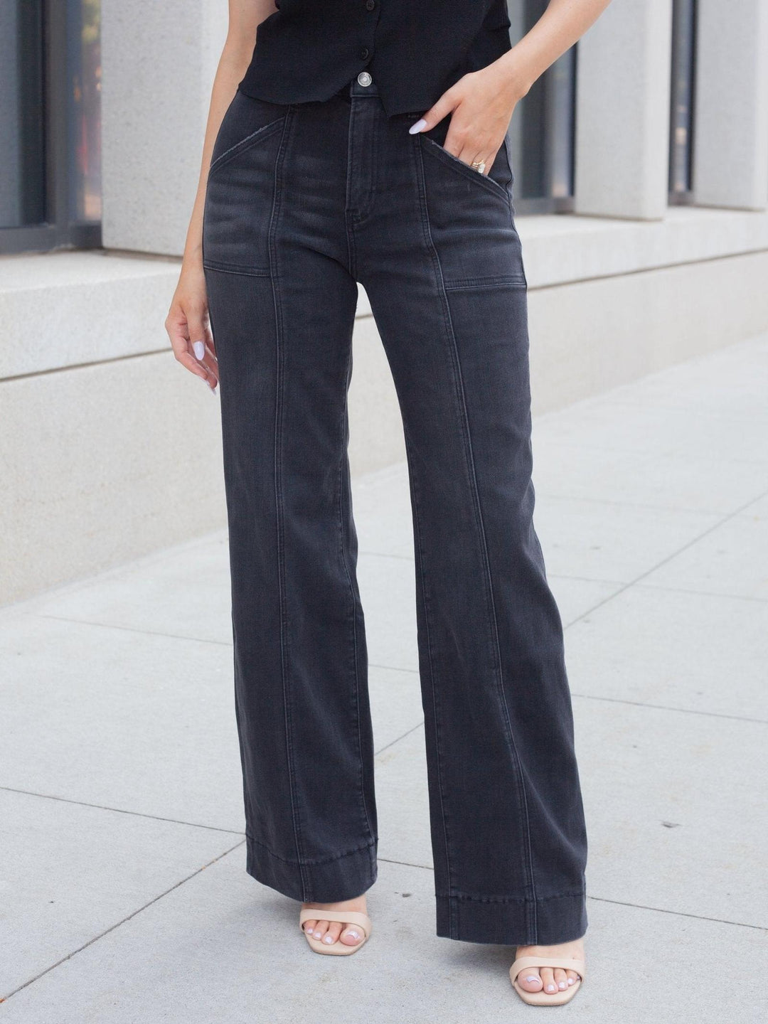 A person wearing dark charcoal wide-leg jeans with a button fly and side pockets, paired with a black top and clear-strap heels, standing on a light concrete sidewalk.
