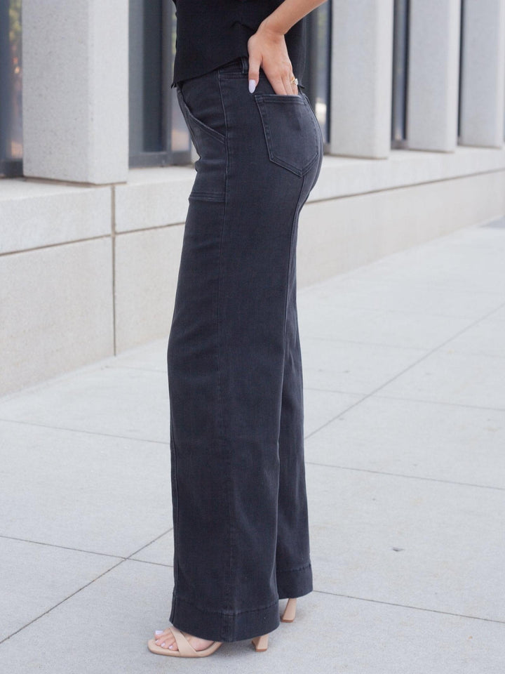 A person wearing dark charcoal wide-leg jeans with a black top, standing outdoors on a light concrete sidewalk in front of a modern building.