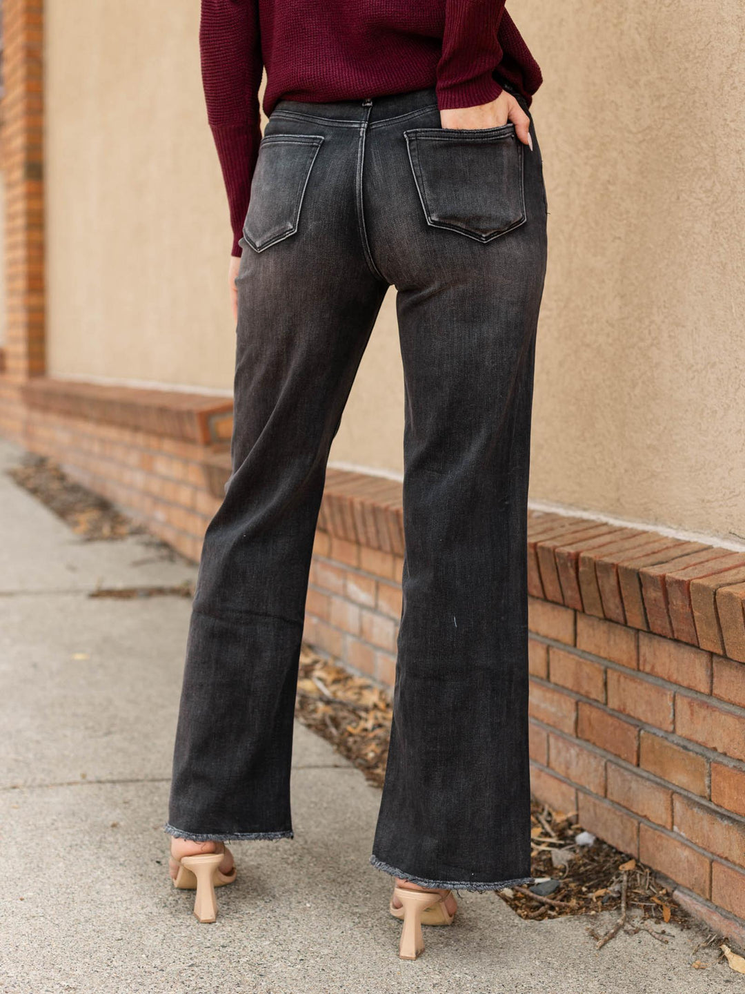 A person shown from the back wearing dark charcoal gray wide-leg flare jeans, a burgundy long-sleeve top, and beige high heels, standing outdoors next to a brick wall and concrete sidewalk.