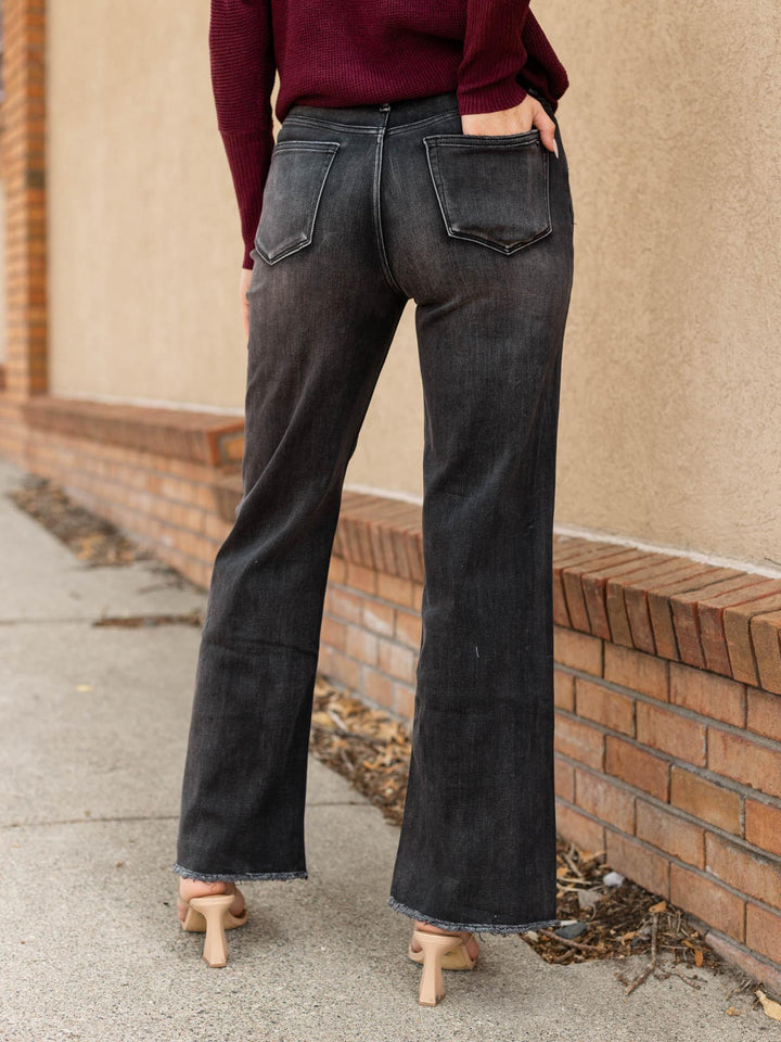 A person shown from the back wearing dark charcoal gray wide-leg flare jeans, a burgundy long-sleeve top, and beige high heels, standing outdoors next to a brick wall and concrete sidewalk.