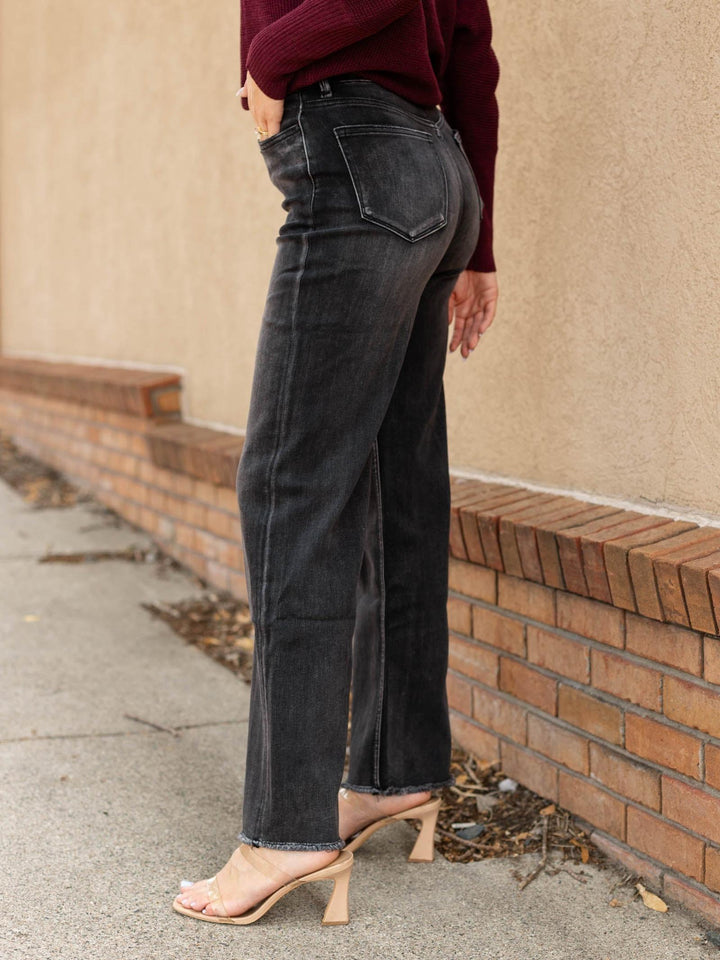 A person wearing dark charcoal gray wide-leg jeans, a burgundy long-sleeve top, and beige heeled sandals, standing outdoors against a beige stucco wall and brick base.