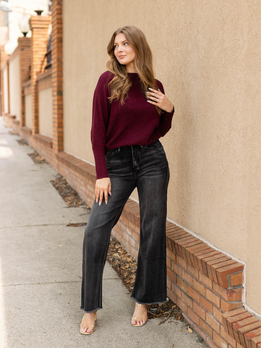 A woman stands barefoot on a sidewalk next to a beige stucco wall and brick base, wearing a long-sleeve burgundy knit top and dark charcoal flared jeans.