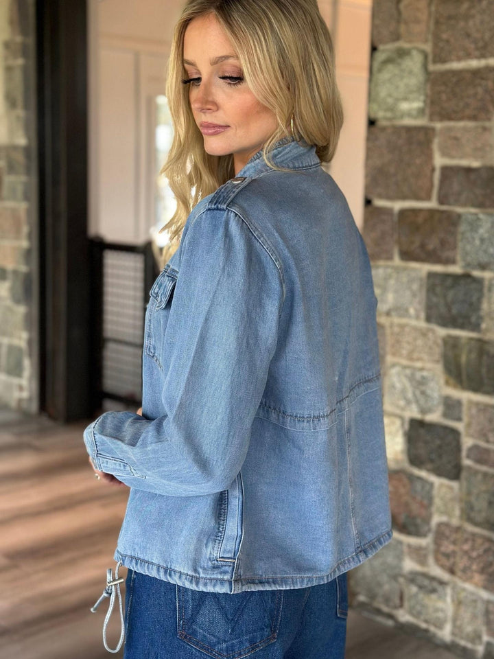 A woman wearing a light blue denim jacket, shown from a side-back angle in an indoor setting with a stone wall background.