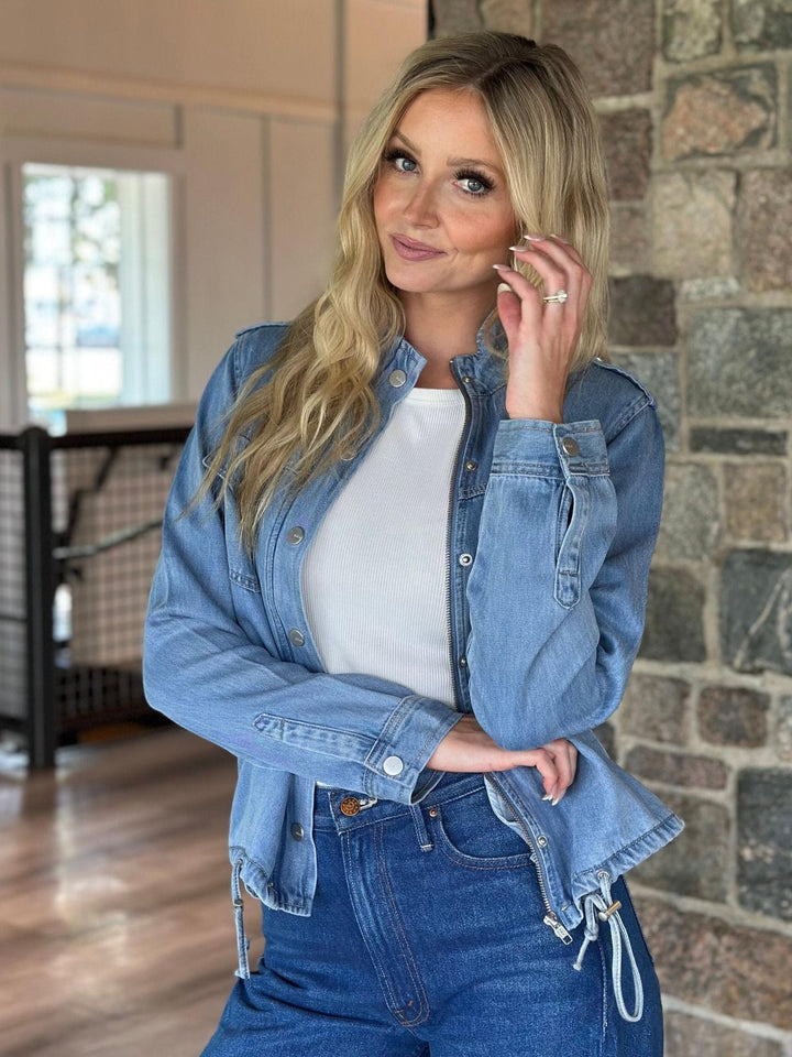 A woman wearing a light-wash blue denim jacket over a white top and dark blue jeans, standing indoors against a stone wall.