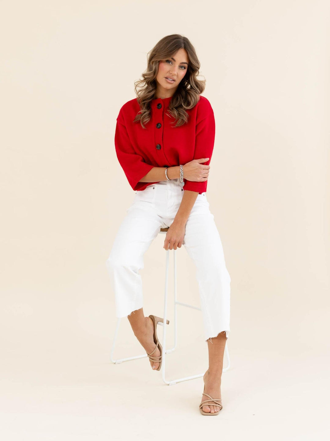 Woman wearing a red top and white pants sitting on a chair against a beige background