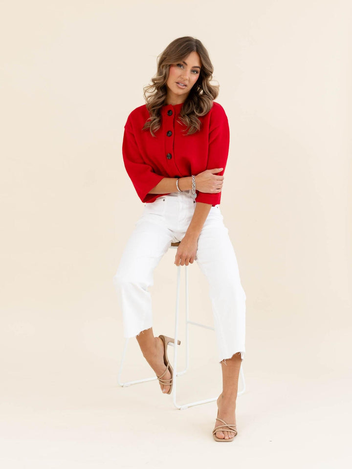 Woman wearing a red top and white pants sitting on a chair against a beige background