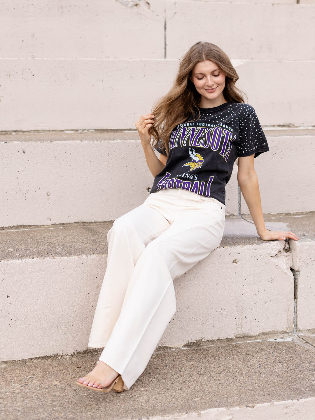 A woman sits on concrete steps wearing a black Minnesota Vikings graphic t-shirt and off-white wide-leg pants.