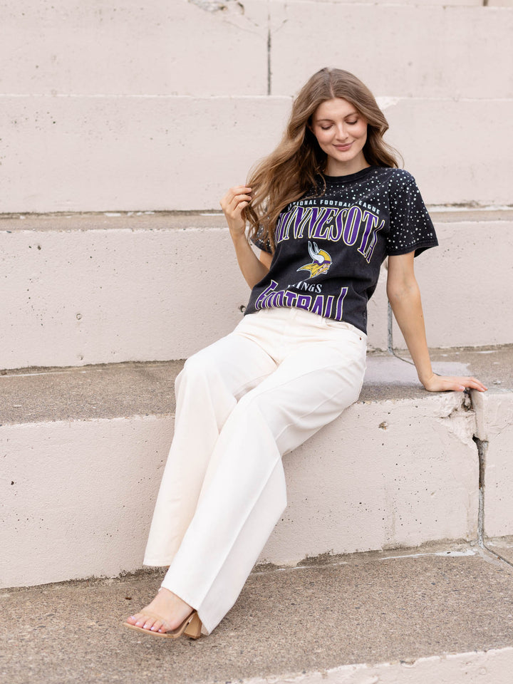 A woman sits on concrete steps wearing a black Minnesota Vikings graphic t-shirt and off-white wide-leg pants.