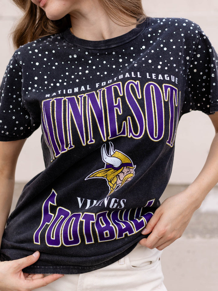 A woman wearing a faded black Minnesota Vikings football graphic t-shirt with purple and white lettering and a white speckled pattern on the sleeves.